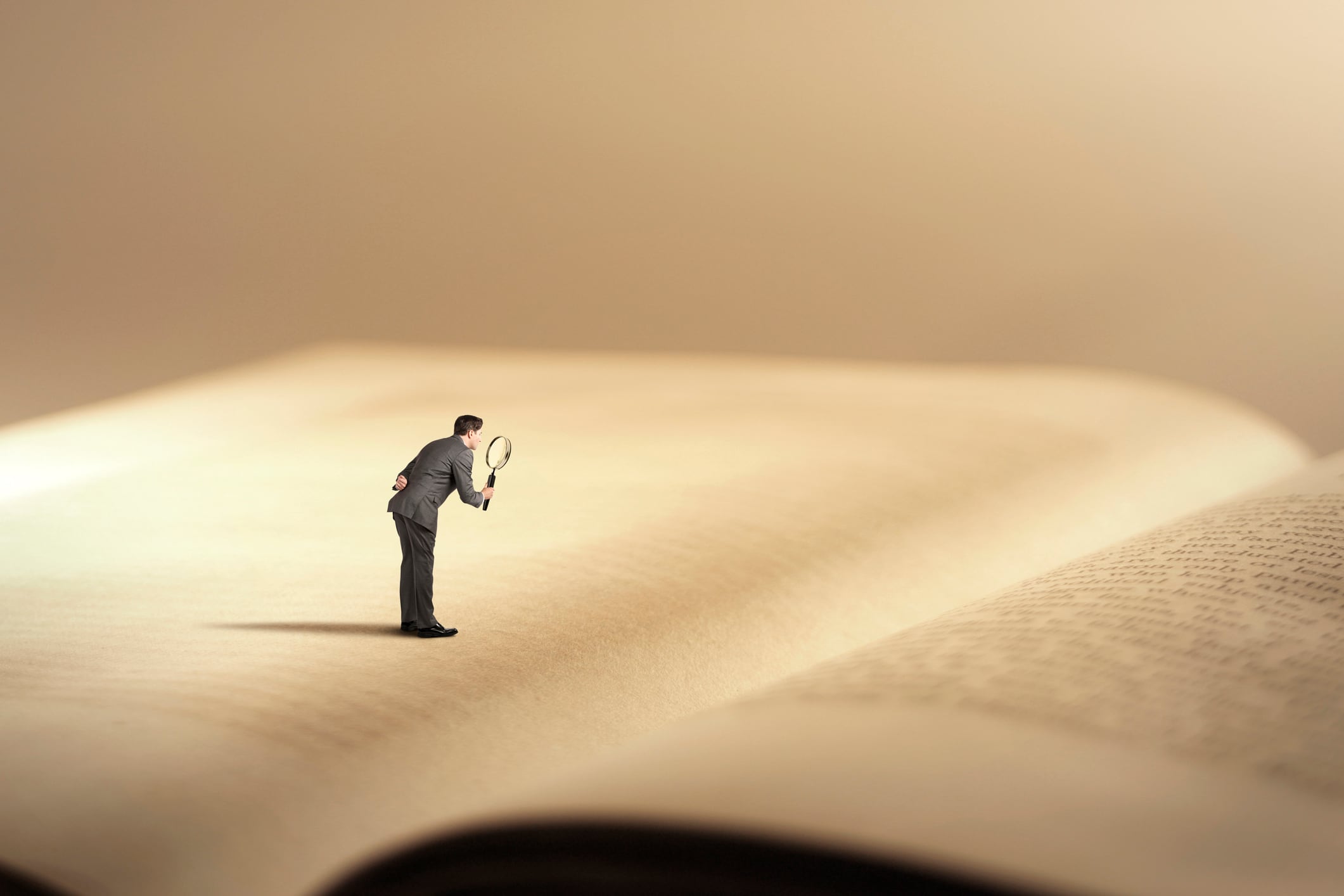 A man stands on a giant book as he looks at it through a magnifying glass.