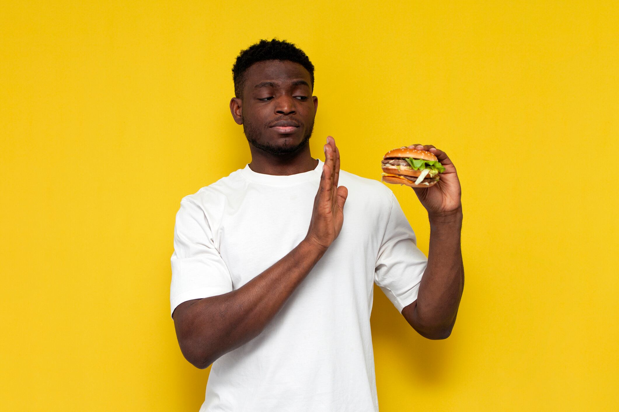 man in white t-shirt refuses burger on yellow isolated background, the guy forbids and says no to fast food