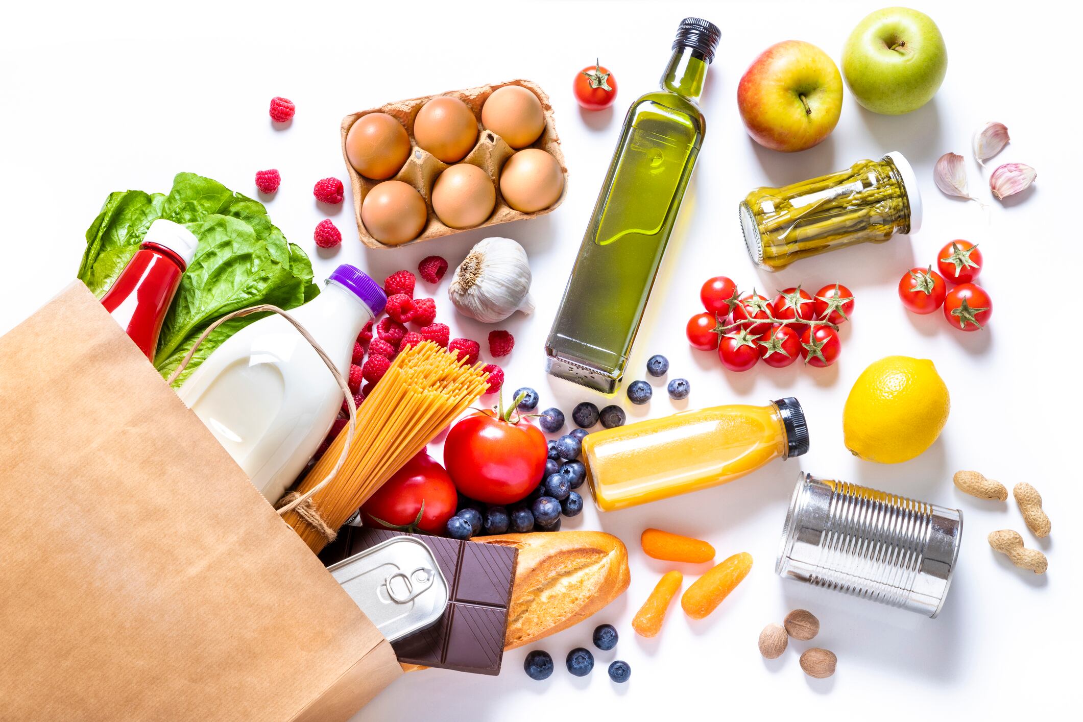 Top view of a paper bag full of canned food, fruits, vegetables, eggs, a milk bottle, berries, mushrooms, nuts, pasta, a chocolate bar and bread. The paper bag is laying on the white background and the food is coming out from it.