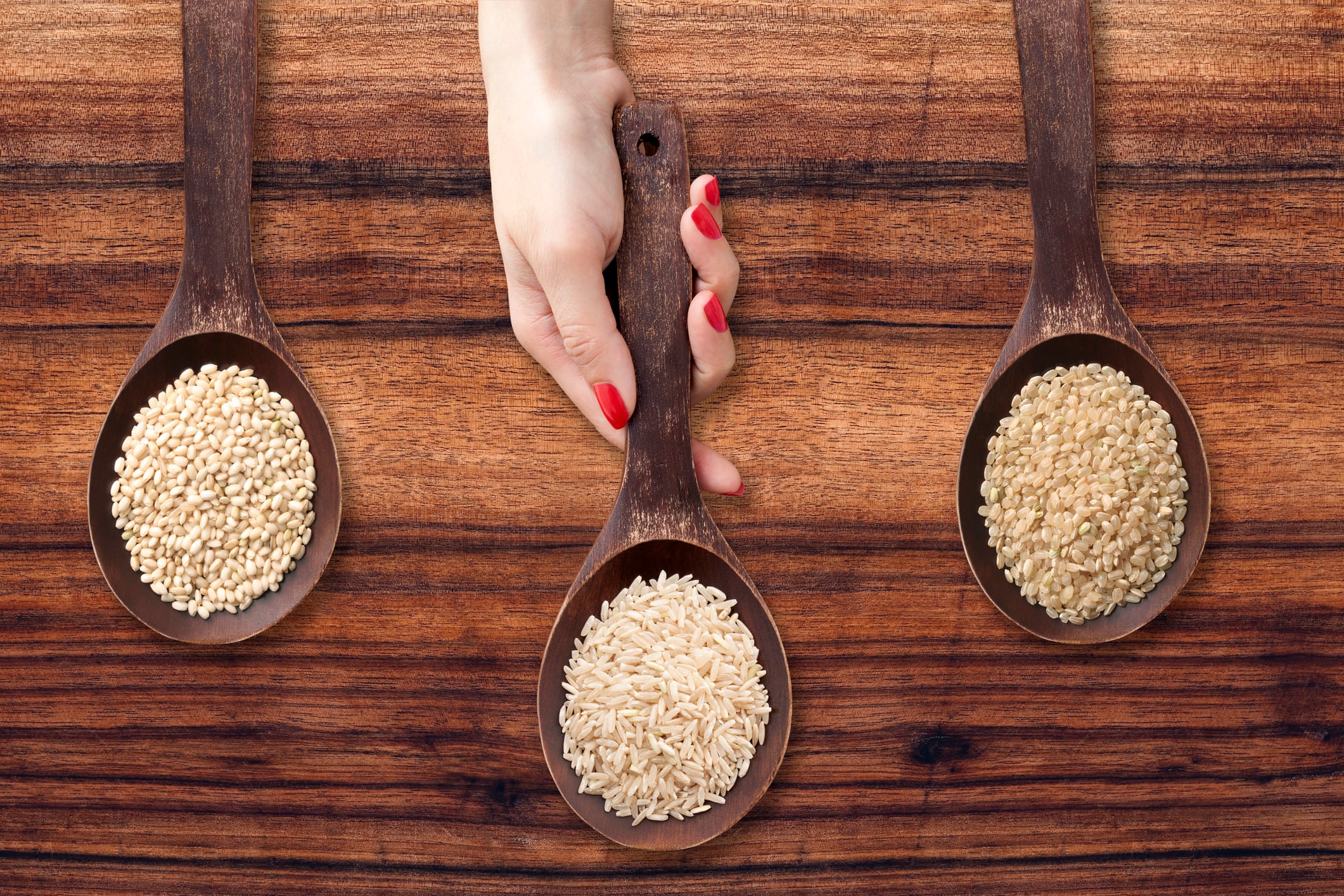 Three wooden spoons with varieties of brown rices and woman hand holding the one in the middle