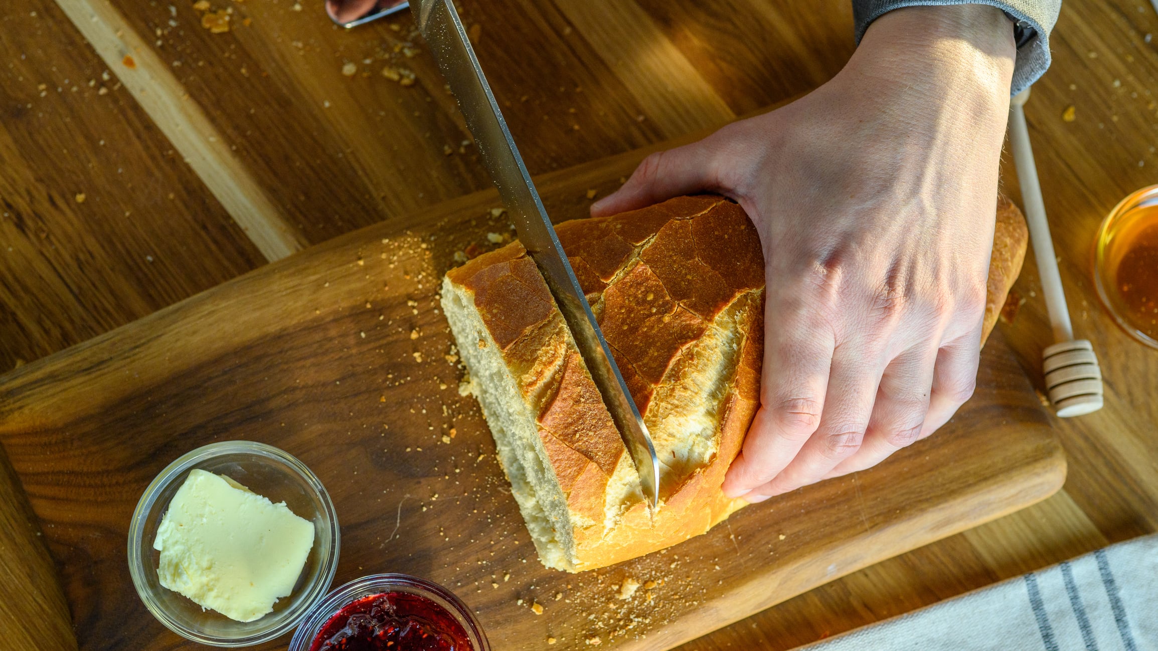 A person expertly slices a crusty loaf of bread on a wooden cutting board, surrounded by jars of spreads, in a sunlit kitchen.