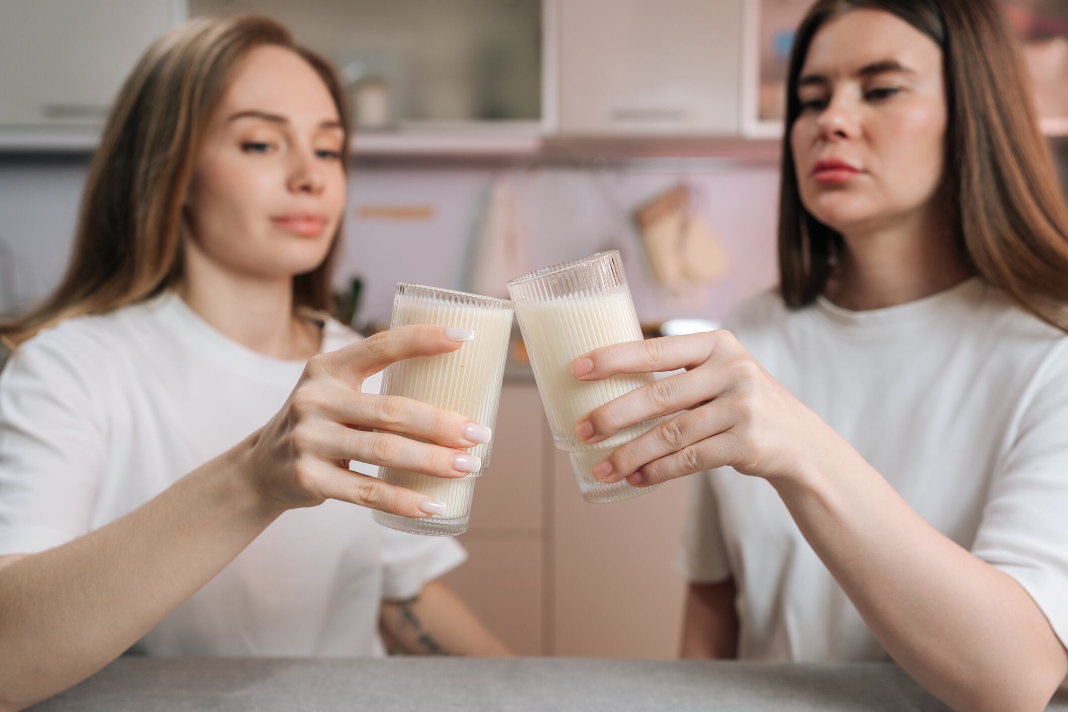 Two young women in white shirts clink glasses of milk or smoothie. Shallow depth of field focuses on hands and drinks. Casual, healthy lifestyle concept in modern kitchen setting.