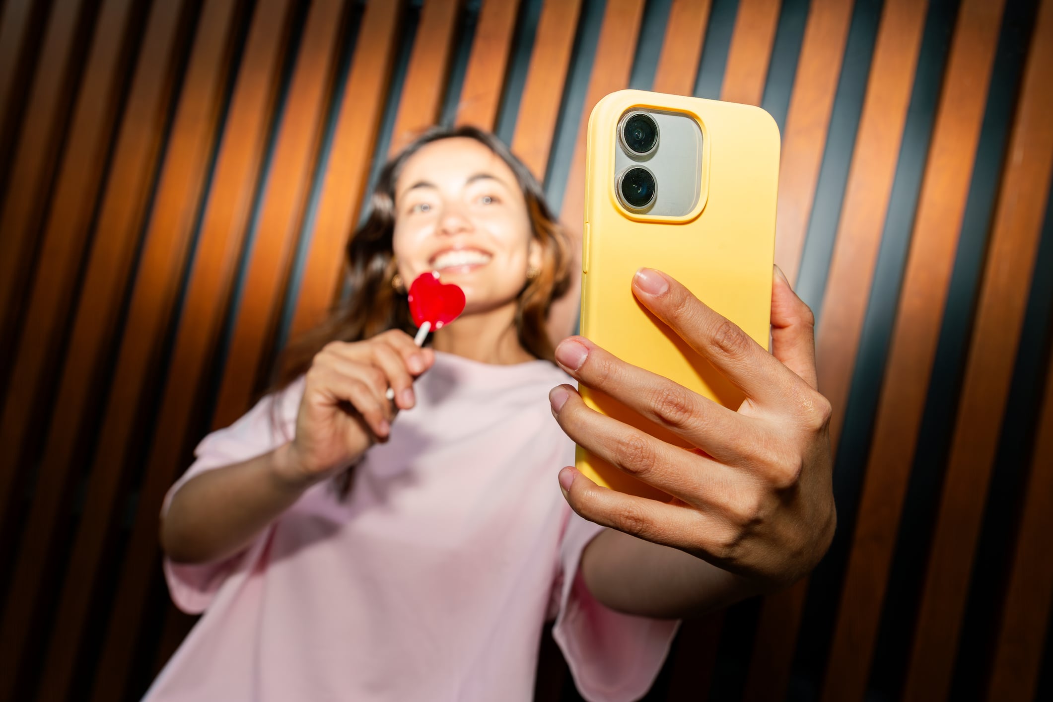 Young adult woman smiling, taking a selfie with her yellow smartphone and a heart lollipop