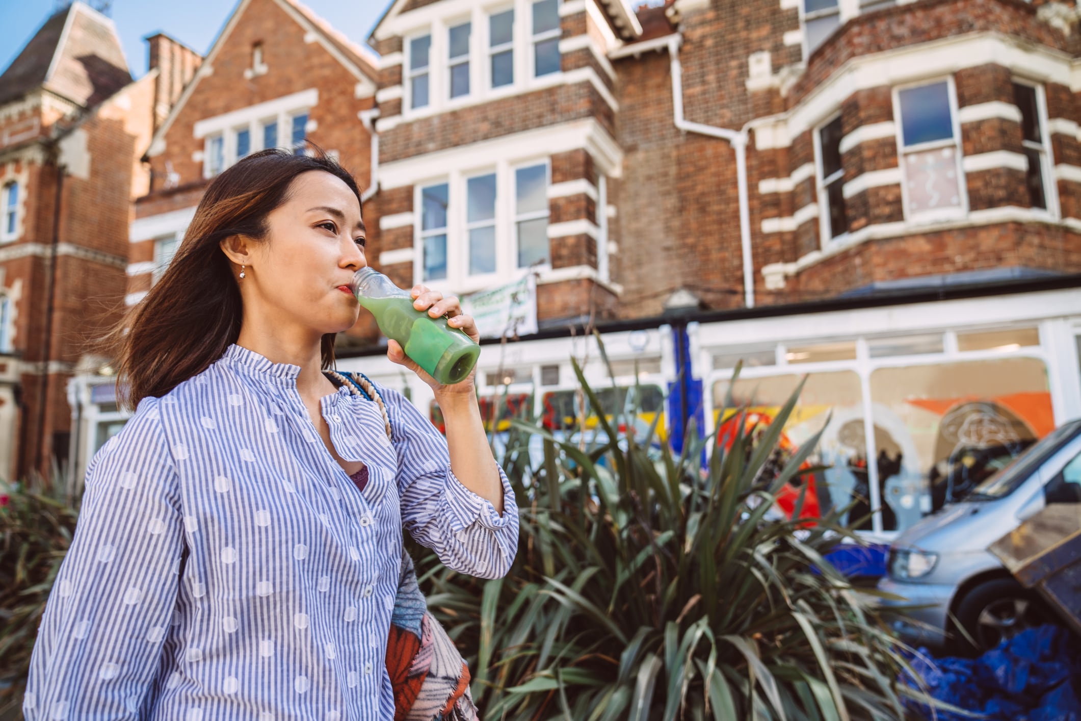 Young cheerful female traveller drinking a bottle of juice while walking on the sidewalk in downtown district.