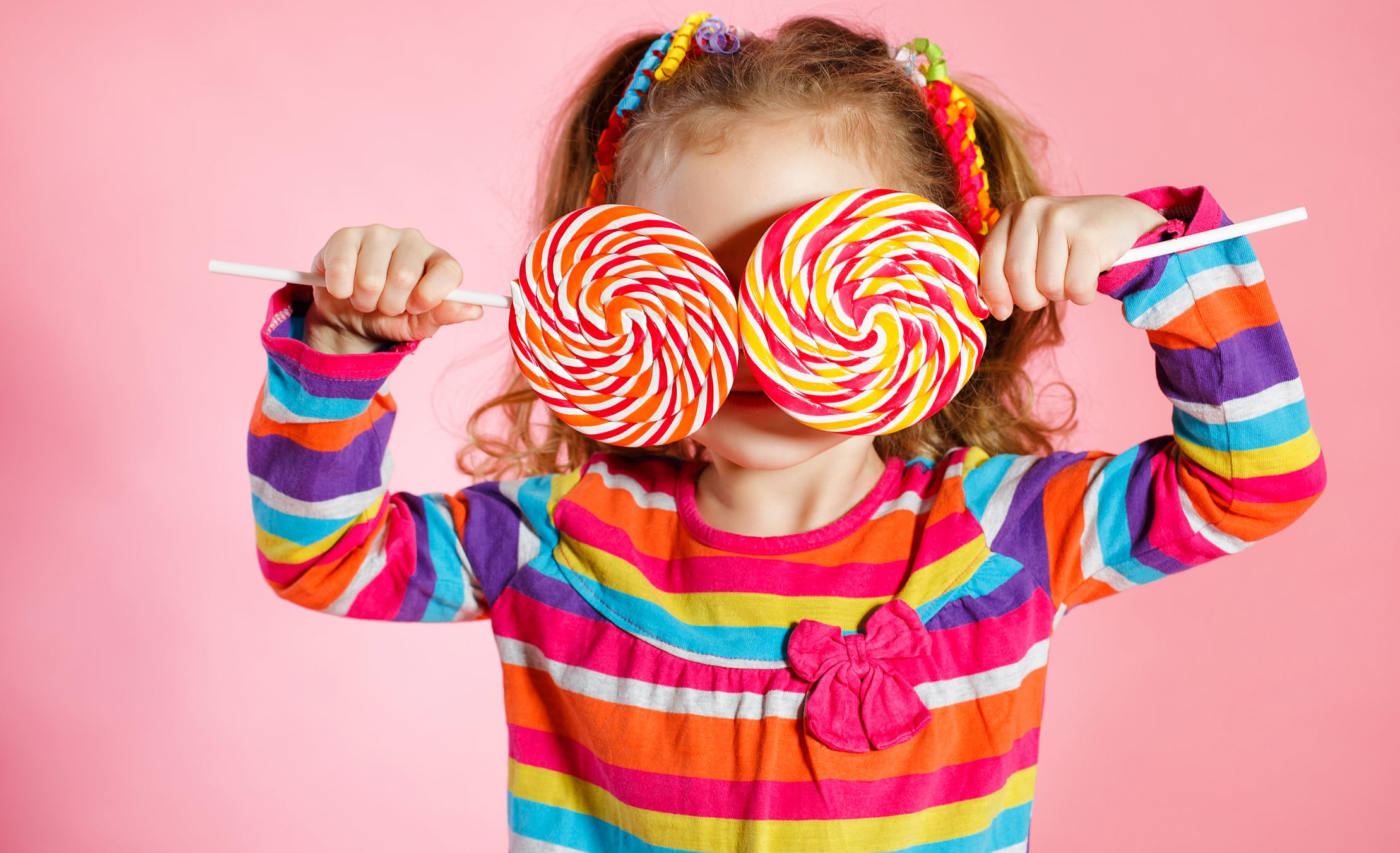 Little girl wearing bright dress, holding two lollipops over her eyes.