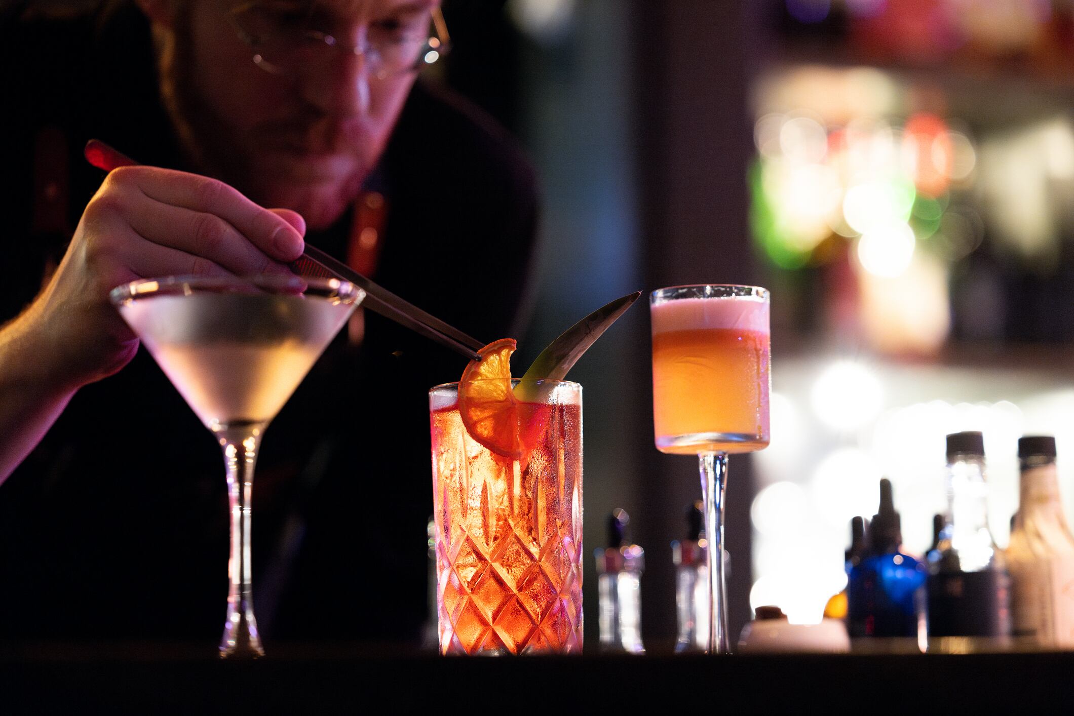 A still photograph of a bartender as he puts an orange slice into a cocktail. The drinks are vibrant and brightly lit, standing out against the low-key lighting of the background. The drinks and bartender's hand are in focus, while the rest remains blurry.