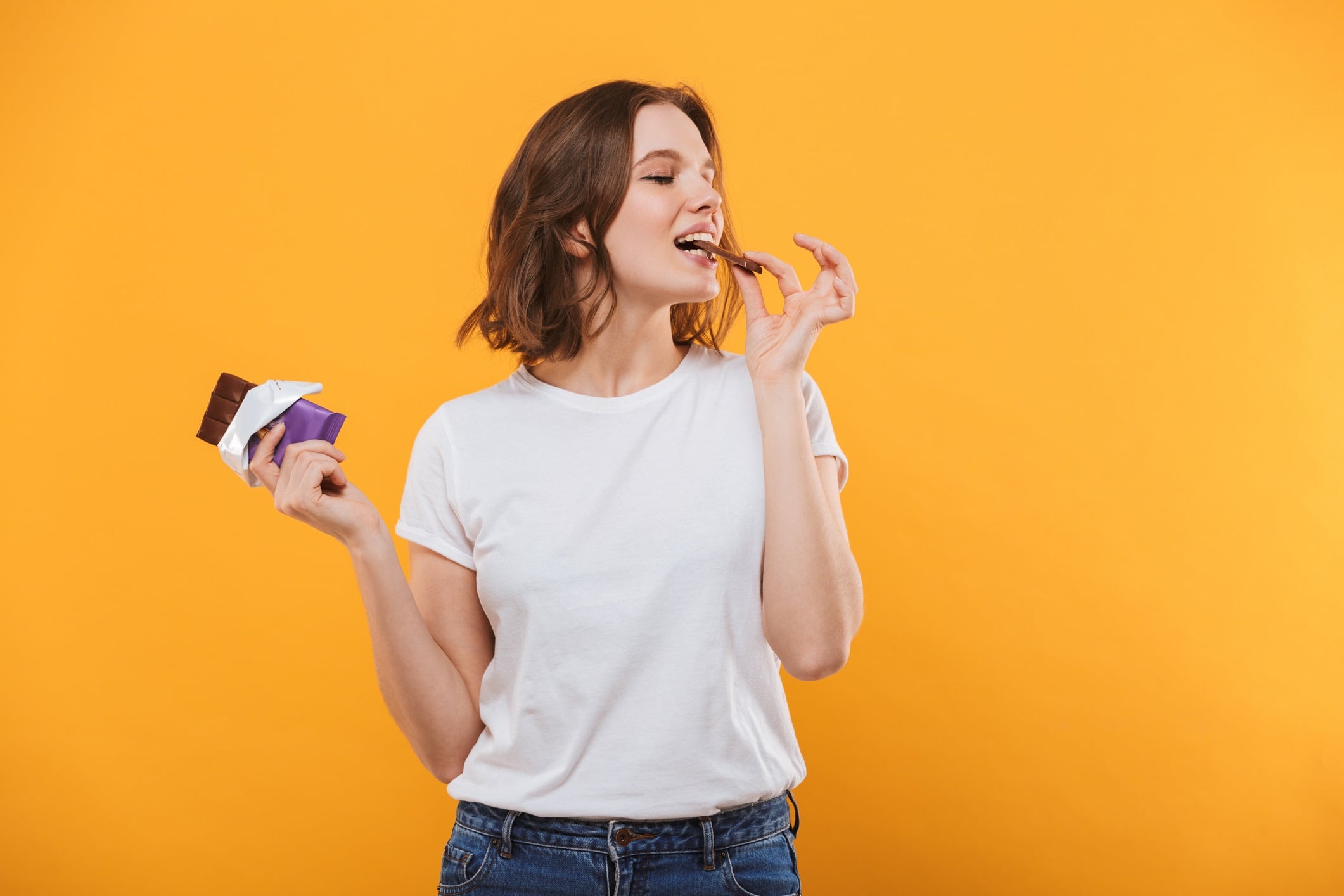 Young woman eating chocolate on yellow background.