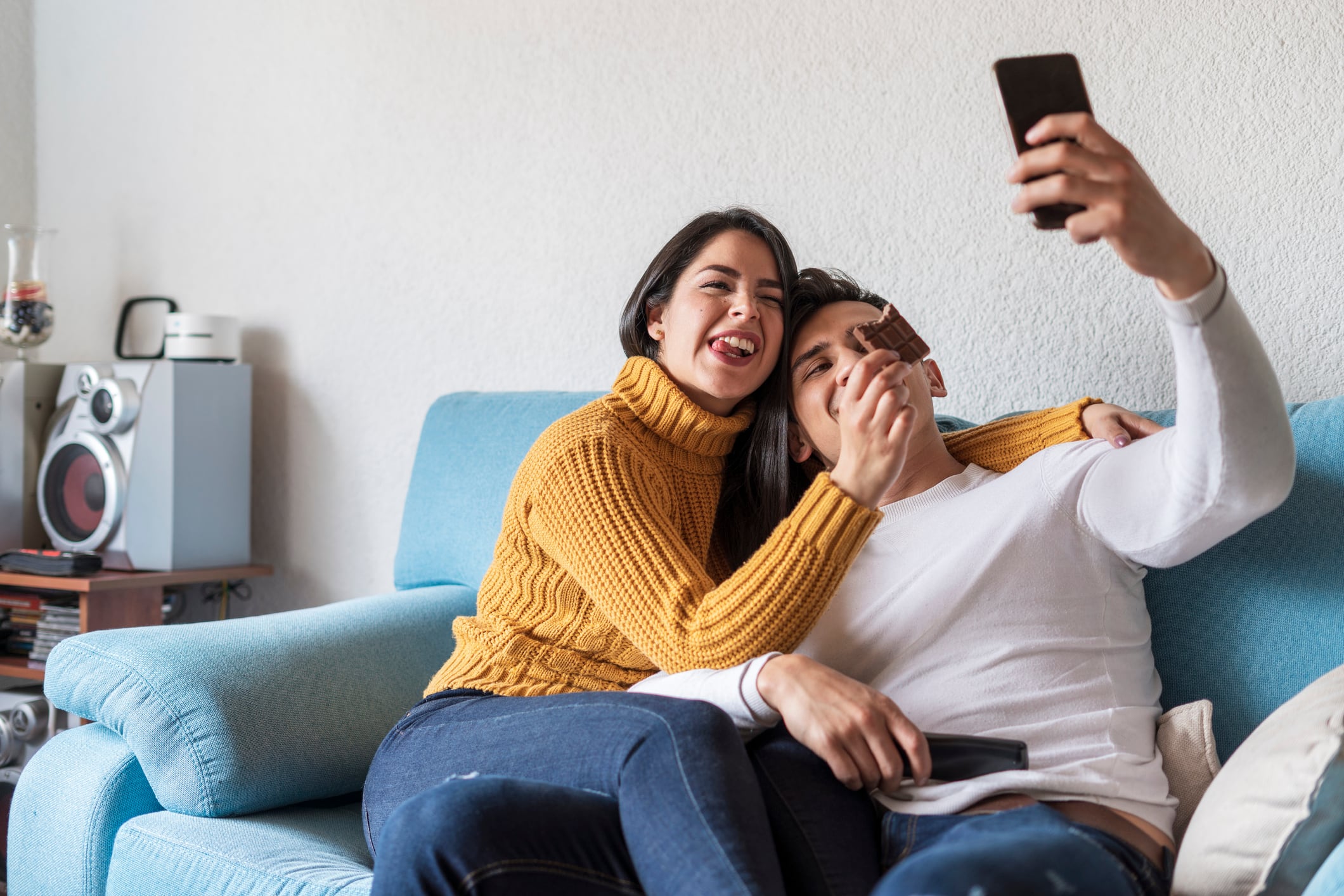 Latin couple of average age of 25 years dressed casually are on the sofa in their living room taking a selfie and eating chocolate.