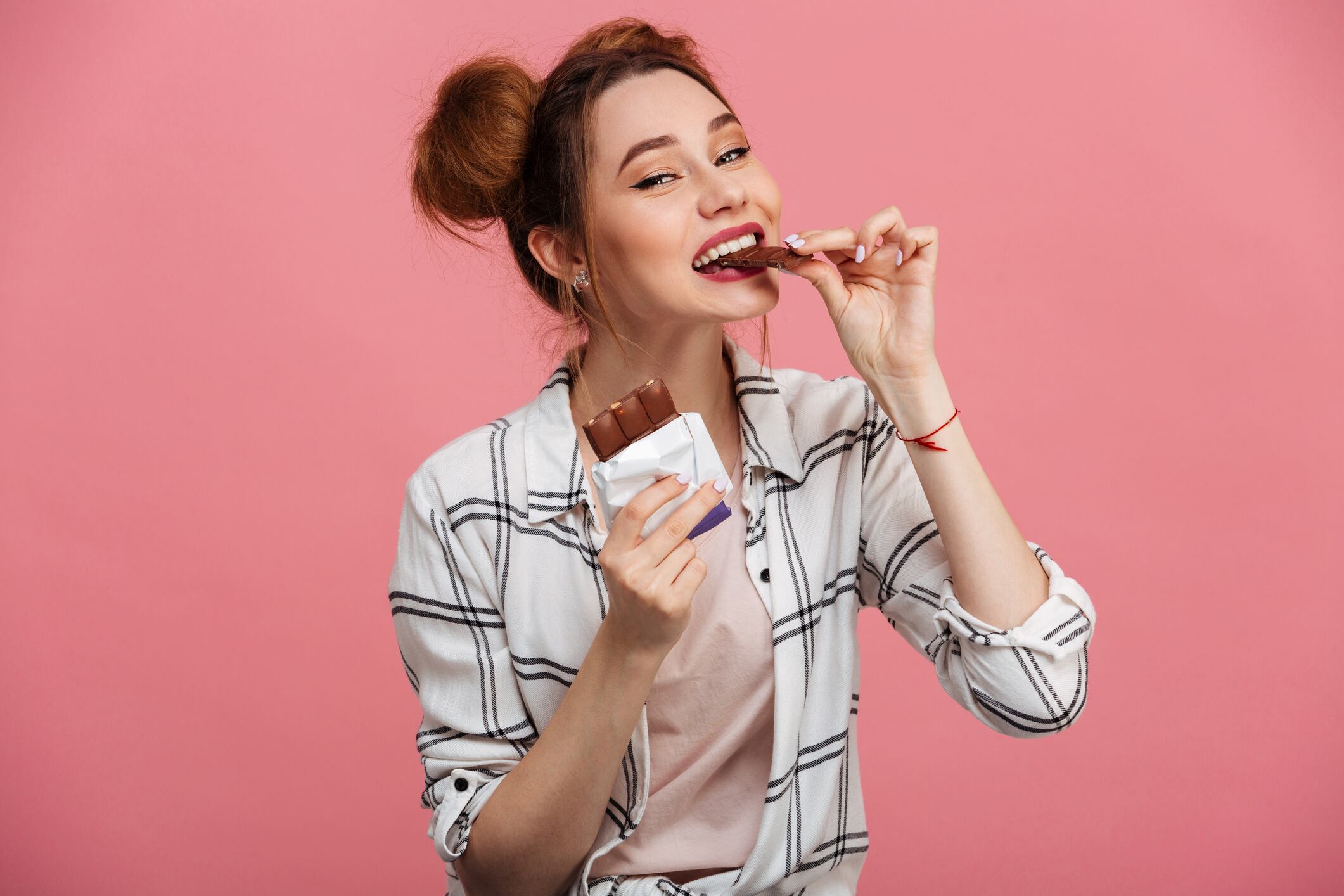 Woman wearing checked shirt, eating chocolate bar, isolated over pink background.