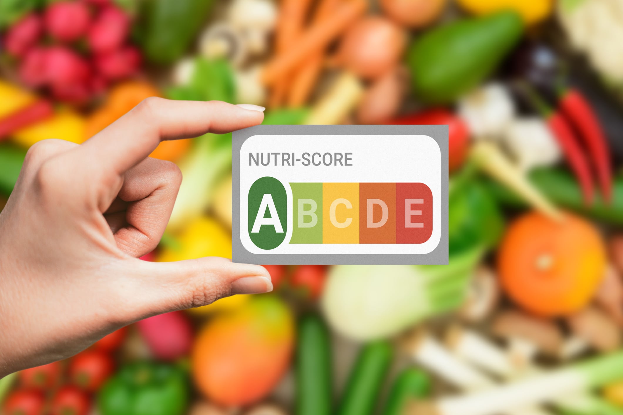 A hand holding a Nutri-Score card indicating nutritional value, with a colorful assortment of healthy foods in the background, symbolizing health, nutrition, and balanced diet.
