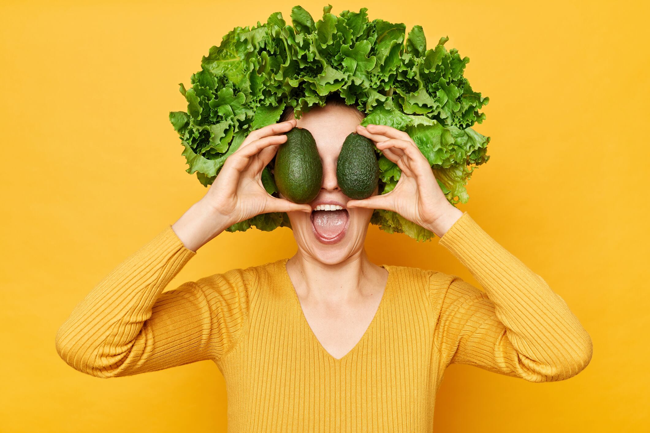 Nutritious food choices. Amazed excited woman with lettuce hair style with green vegetables on her head isolated over yellow background covering eyes with avocado screaming wow.